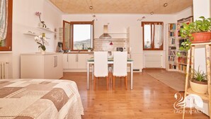 A spacious room with a bed, a dining area with chairs and a table, a kitchen with white cabinets, and a bookshelf filled with books. - Appartamento Il Moresine (Roe Volciano)