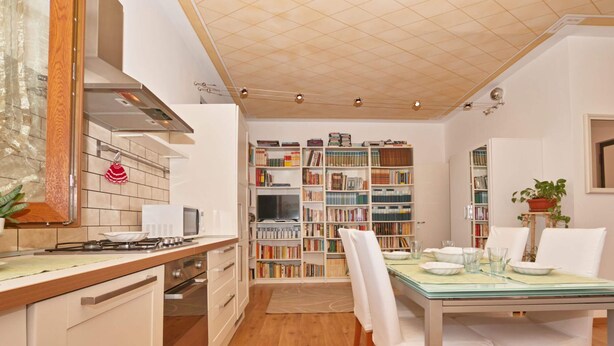 A compact kitchen with a dining area, featuring a built-in bookshelf filled with books, a gas stove, and a dining table set for two. - Appartamento Il Moresine (Roe Volciano)