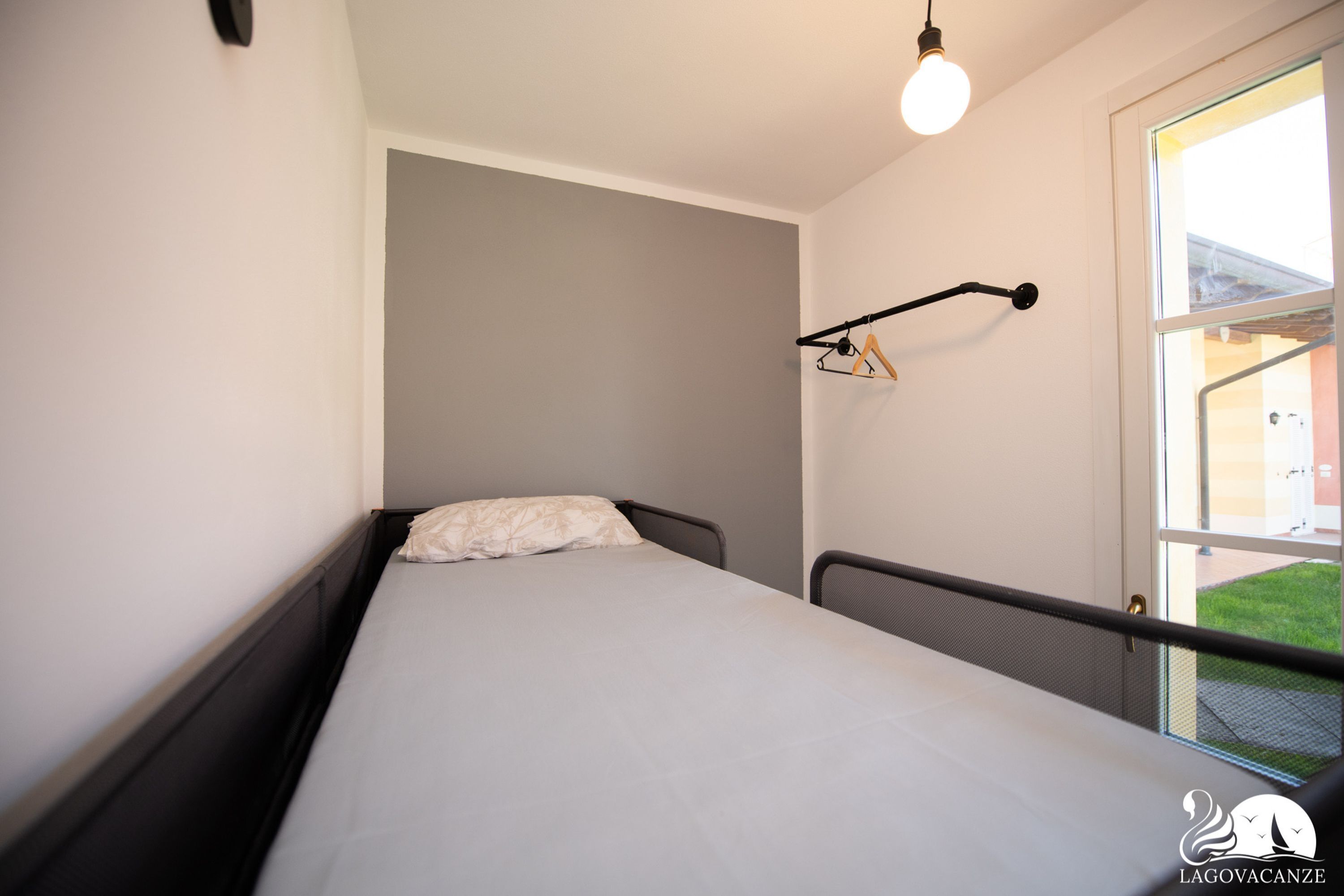 A small, single-bed room with a window, a hanging light fixture, and a coat rack.