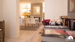 Uma sala de estar moderna com uma mesa de centro de madeira, uma área de jantar com mesa e cadeiras de vidro, e uma estante com livros e objetos decorativos.