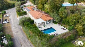 Uma casa moderna com piscina, cercada por vegetação exuberante e uma estrada com carros estacionados.