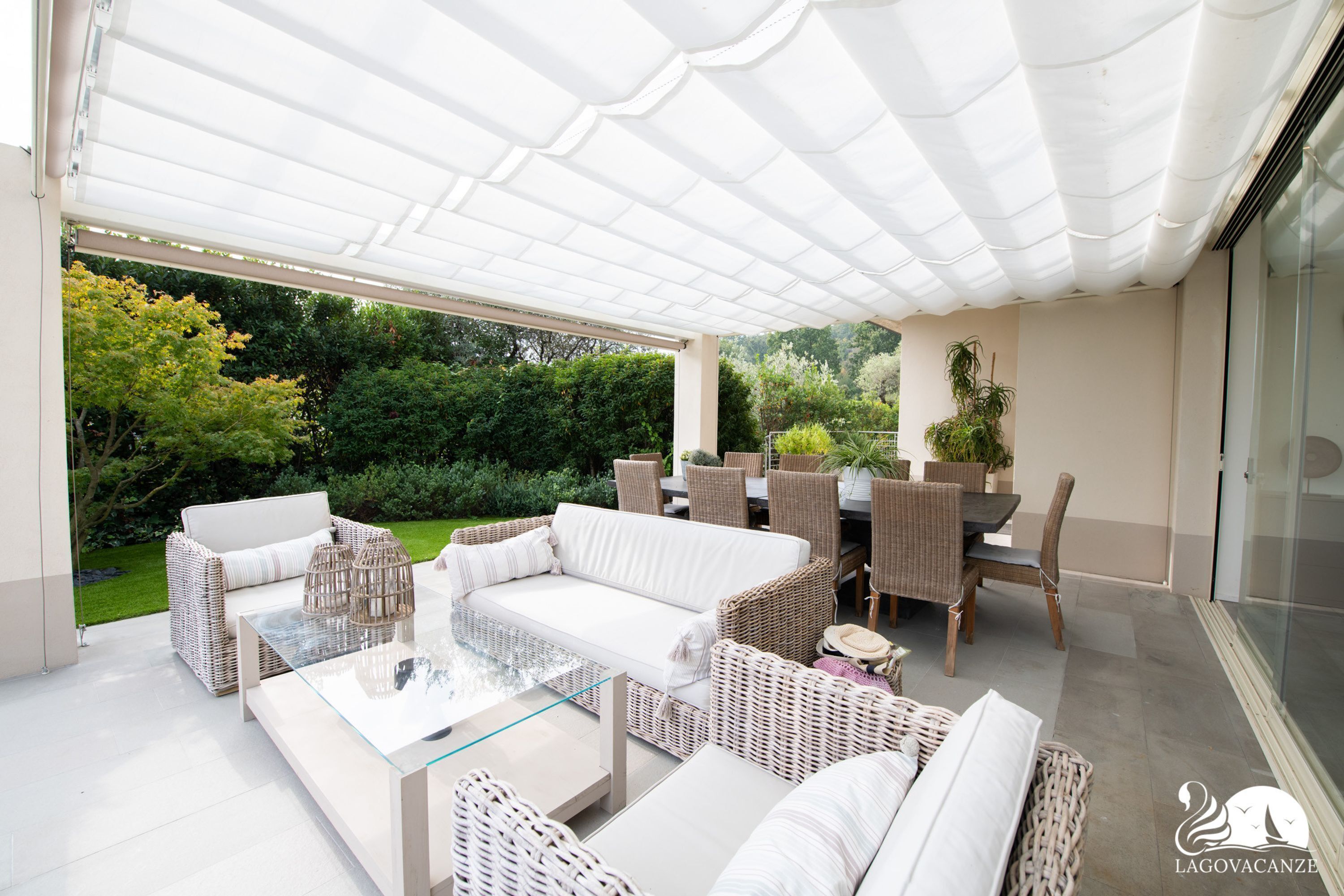 A covered outdoor seating area with wicker furniture, a glass-top table, and a view of the garden.