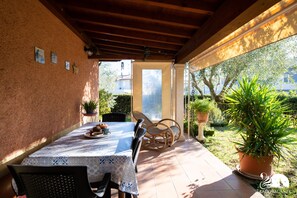 Un patio cubierto con una mesa dispuesta para dos personas, rodeado de plantas en macetas y vistas del entorno.