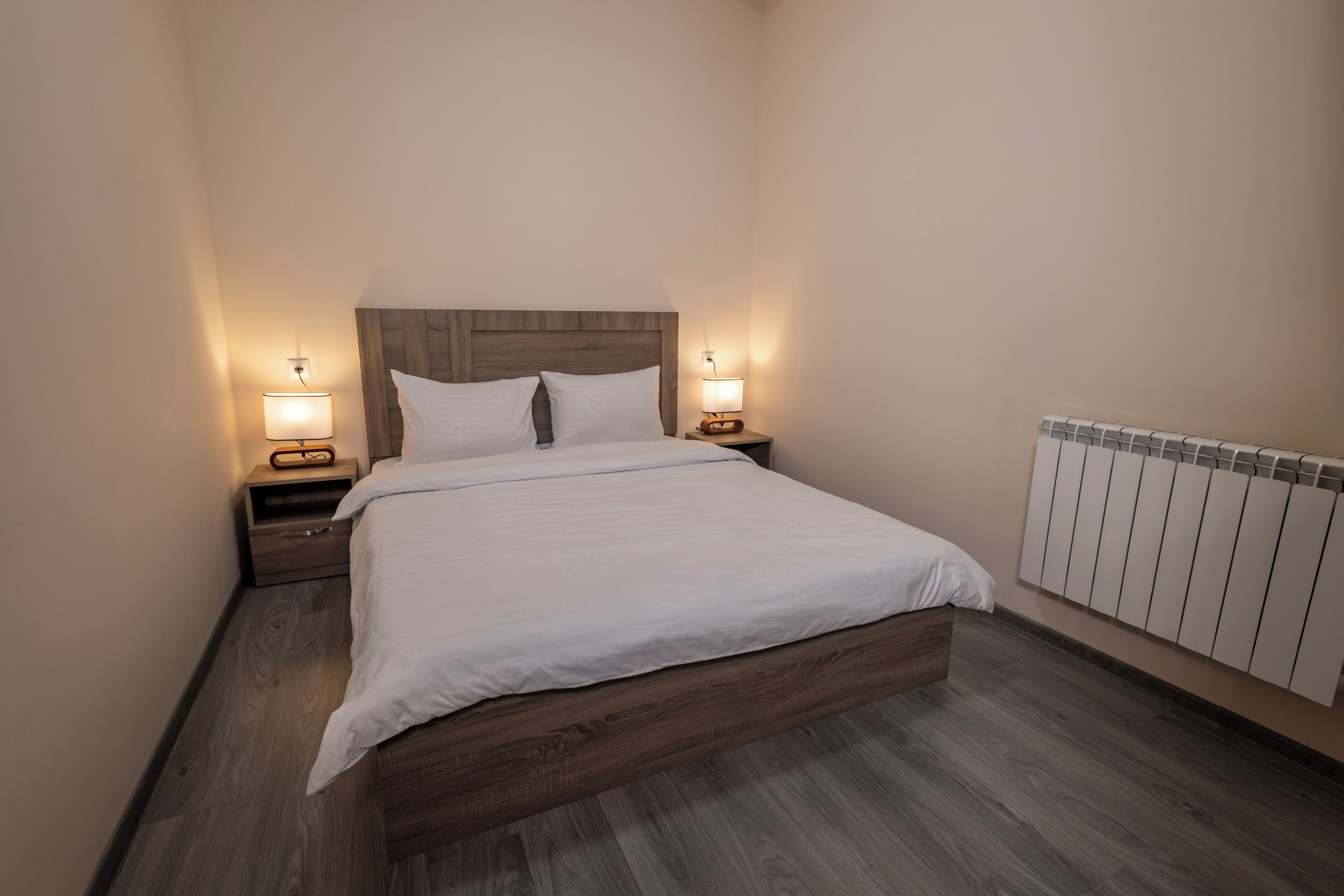 A neatly made bed with a wooden headboard, two bedside tables with lamps, and a radiator on the wall.