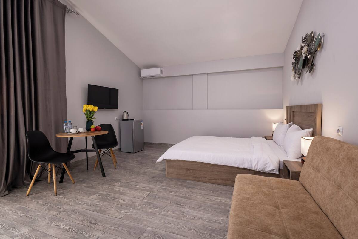 A hotel room with a bed, a sofa, a desk with chairs, a TV, and a small kitchenette.