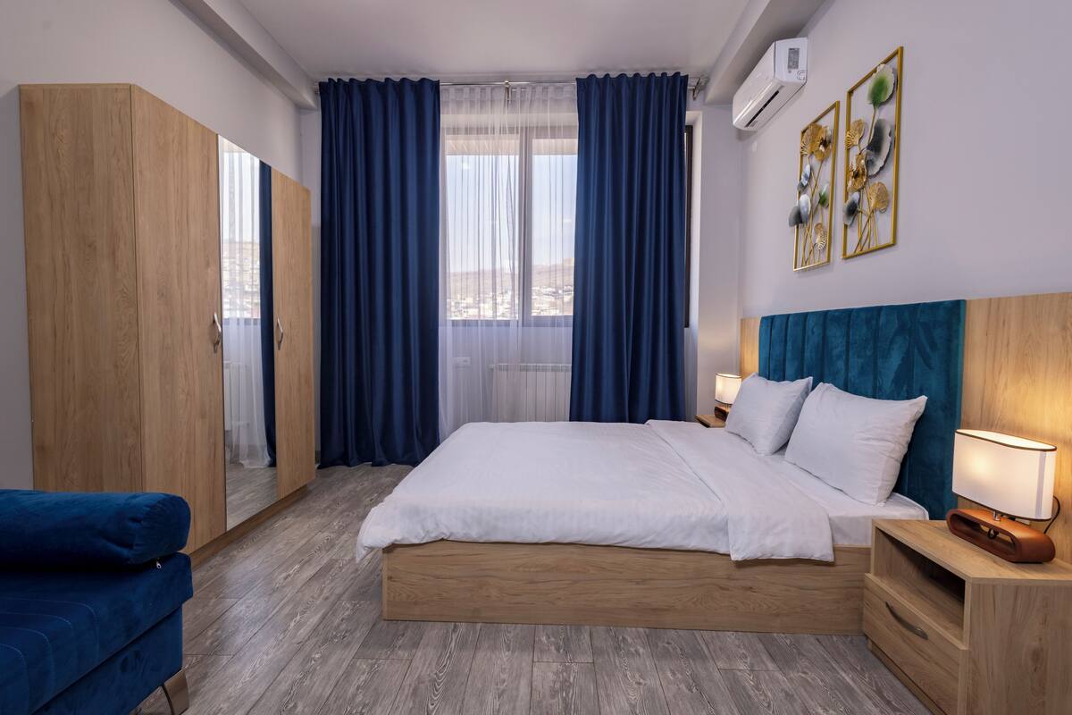 A bedroom with a large bed, a wardrobe, a nightstand with a lamp, and a view through the window.