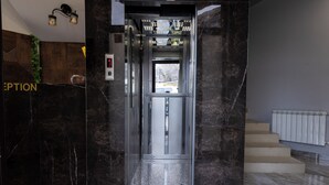 An elevator with a glass door, a reception area with a wooden wall and a plant, and a staircase with a radiator. - Horizon Hotel Yerevan (Yerevan)