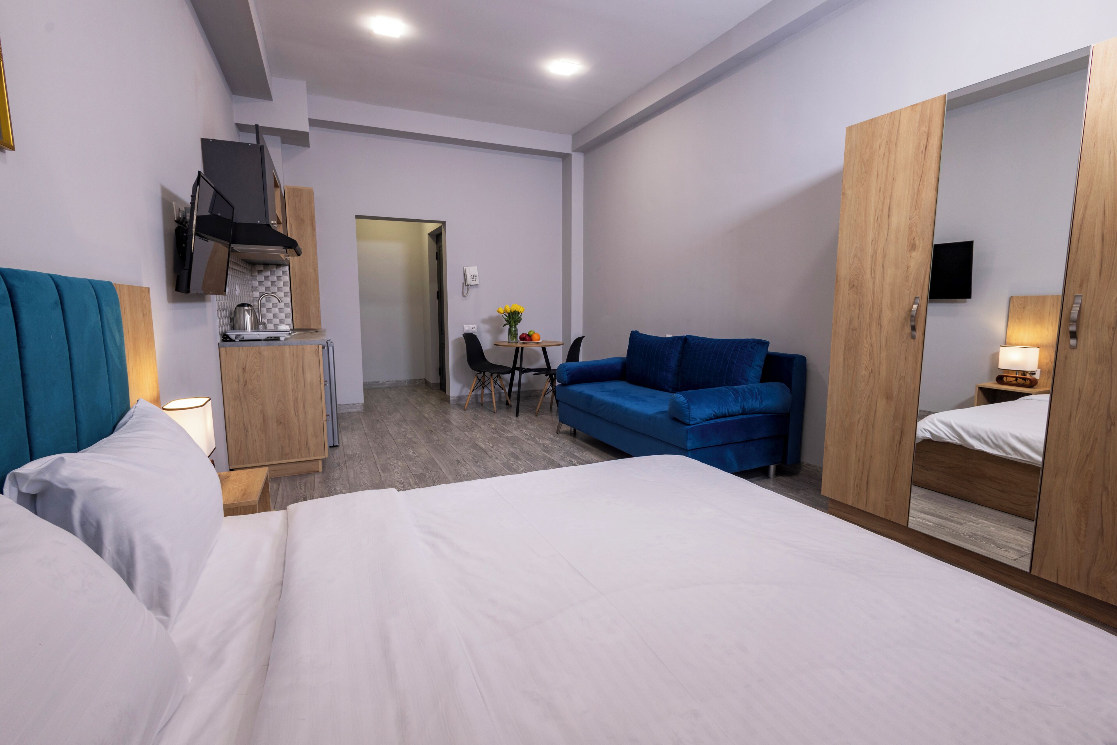A hotel room with a bed, a blue sofa, a small table with a vase, a kitchenette, and a wardrobe with a mirror.