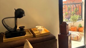 Ein Zimmer mit einer Couch, einem Schreibtisch mit Lampe, einem Tablett mit Tassen und Blick auf Gebäude und einen Baum.