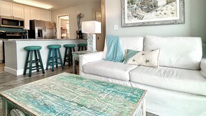 A modern kitchen with white cabinets, a central island with stools, a dining area with a white sofa, a coffee table, and a framed beach scene on the wall. - Surfside #11, Beautiful Beachfront Luxury Condo,  Allergy Friendly:No Pets. (Mexico Beach)