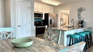 A kitchen with white cabinets, a wooden table with a green bowl, and teal barstools. - Surfside #11, Beautiful Beachfront Luxury Condo,  Allergy Friendly:No Pets. (Mexico Beach)