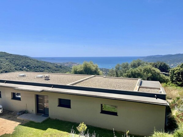 Ein modernes Gebäude mit flachem Dach, kleinem Eingang und Blick auf Meer und Hügel.
