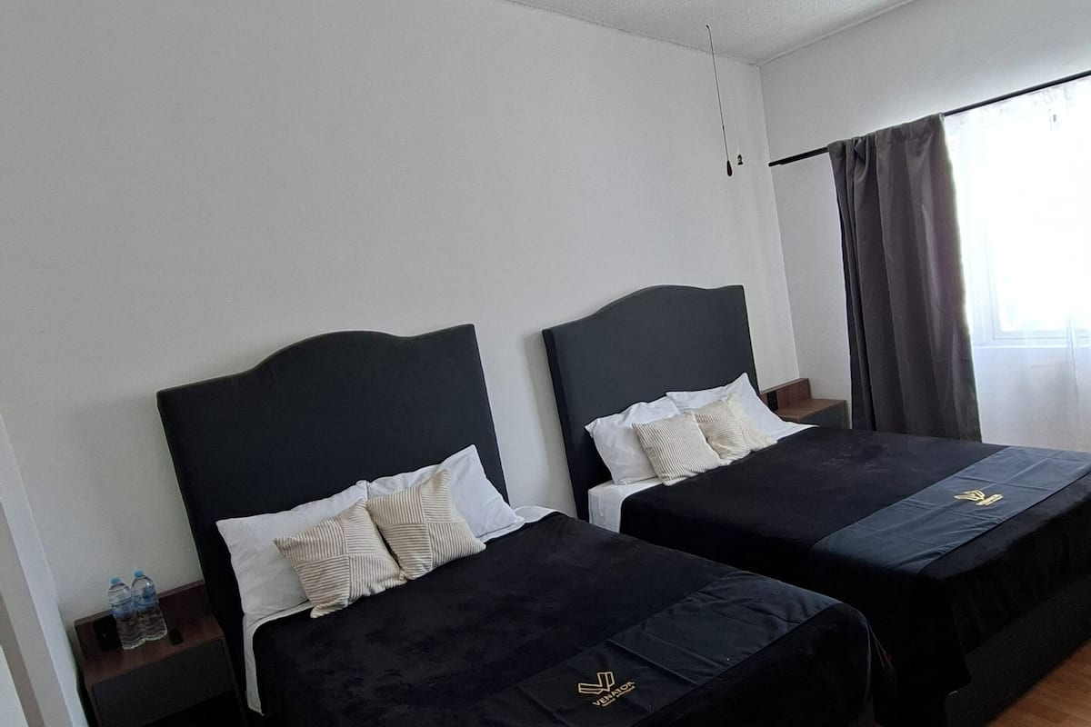 A hotel room with two beds, a ceiling fan, wooden flooring, and a window with curtains.