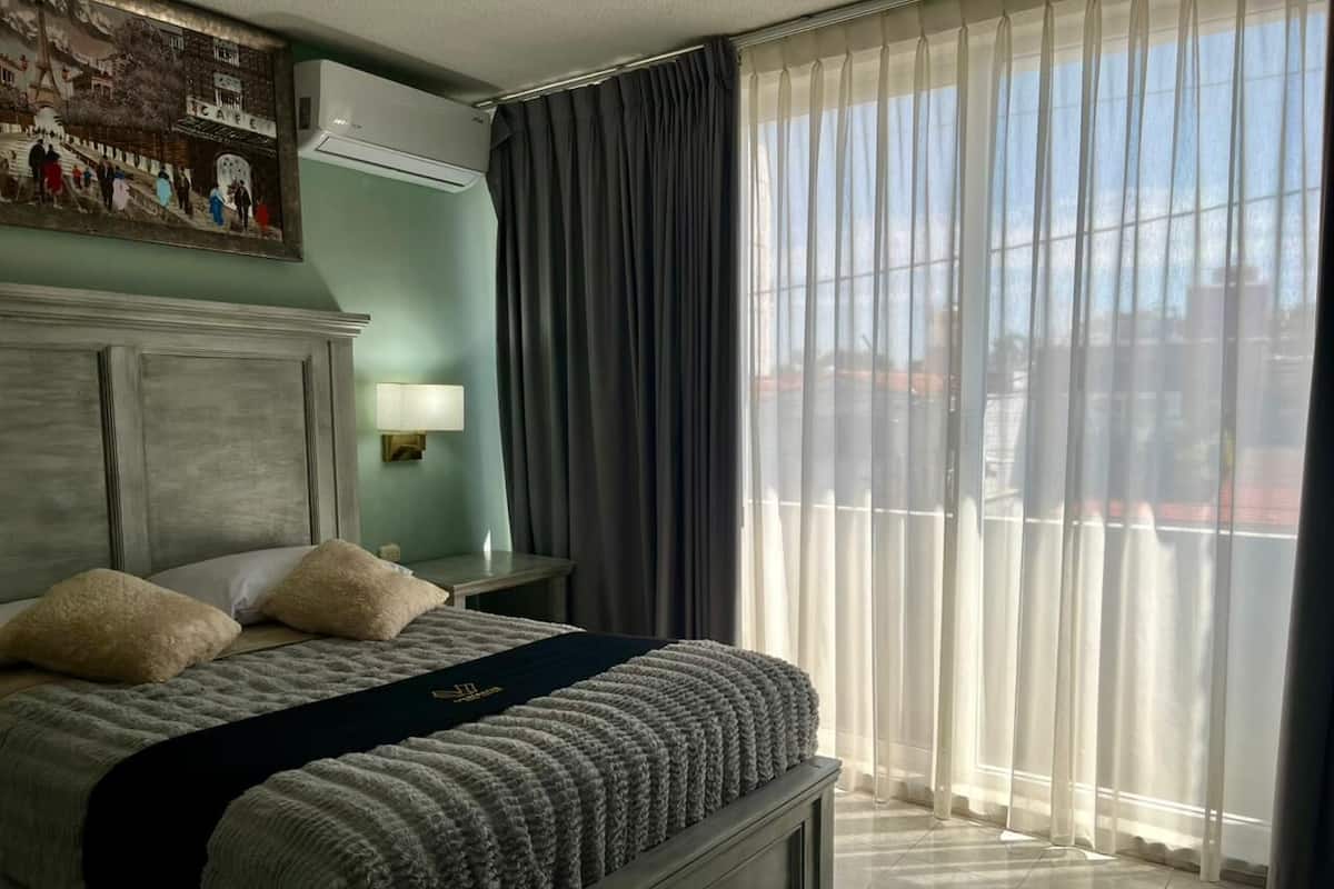 A hotel room with a large bed, a nightstand, a lamp, and a view through sheer curtains.
