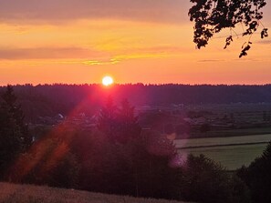 A sunset over a rural landscape with a forested hill and a small village in the distance.