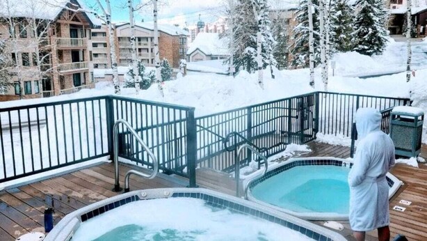 A hot tub area with a wooden deck and metal railings, surrounded by snow-covered trees and buildings. - Large Luxury: Fun 4 the Whole Family. 2 min walk to Slopes. Hot Tubs. Pool Table (Steamboat Springs)