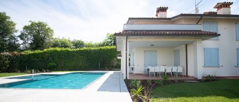 Una casa de dos pisos con piscina, un patio cubierto con mobiliario exterior y un jardín bien cuidado.