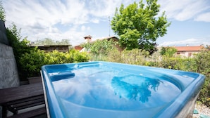 Ein Whirlpool in einem Garten mit einer hölzernen Bank, umgeben von viel Grün und Häusern im Hintergrund.