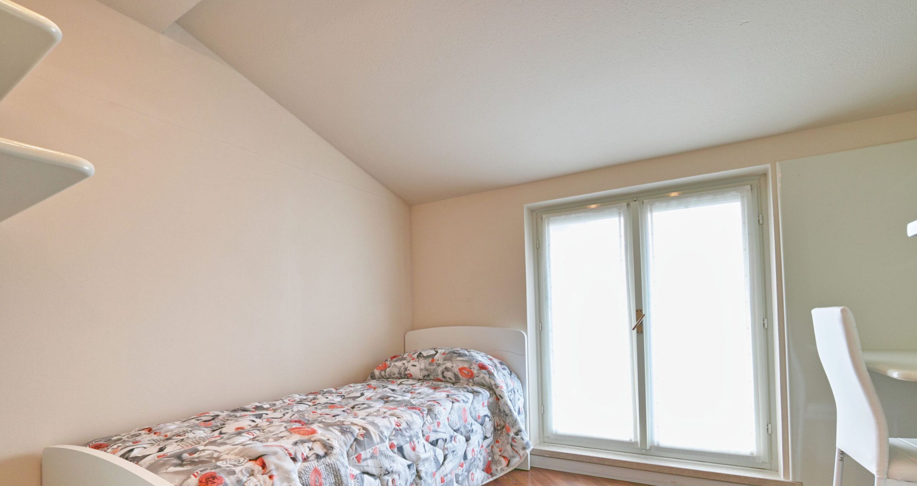 A small, single-bed room with a window, a chair, and a white wall.