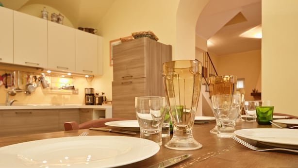 Ein Esstisch ist gedeckt mit Tellern, Besteck und klaren Gläsern in einer hellen Küche, im Hintergrund sind weiße Schränke und ein Holzschrank zu sehen.