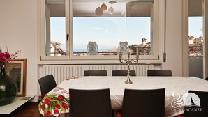 Uma sala de jantar com mesa posta para seis pessoas, um vaso com flores e vista para a paisagem costeira através de uma grande janela.