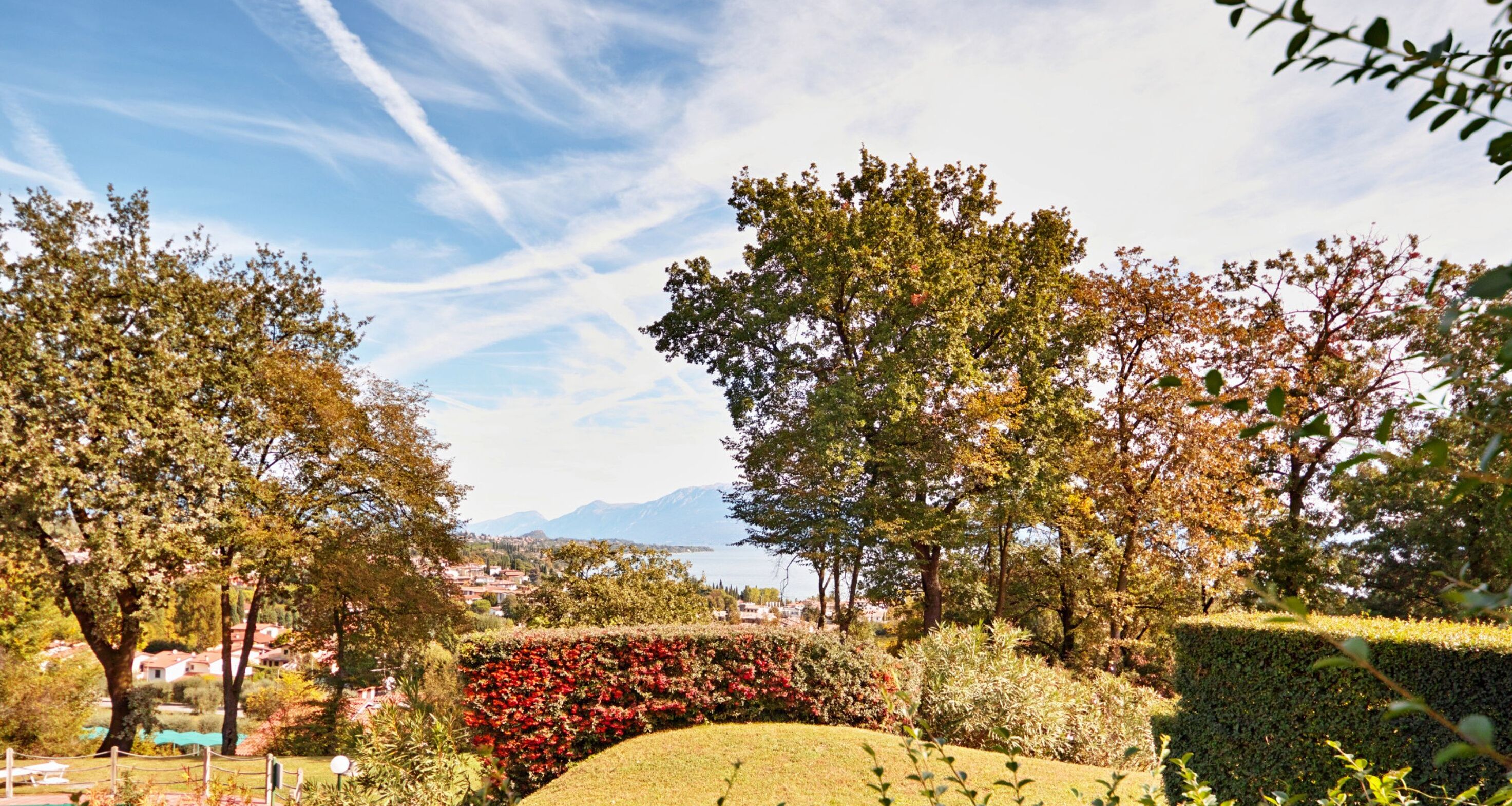 A landscaped garden with various trees and shrubs, a well-maintained lawn, and a distant view of a lake and mountains.