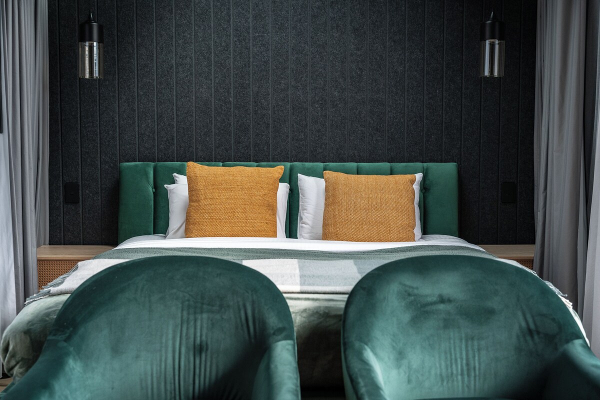 A bed with green upholstered headboard and pillows, a dark textured wall, and two pendant lights.