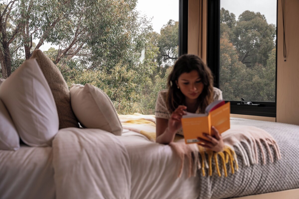 A person reading a book on a bed with a view of trees through a window.