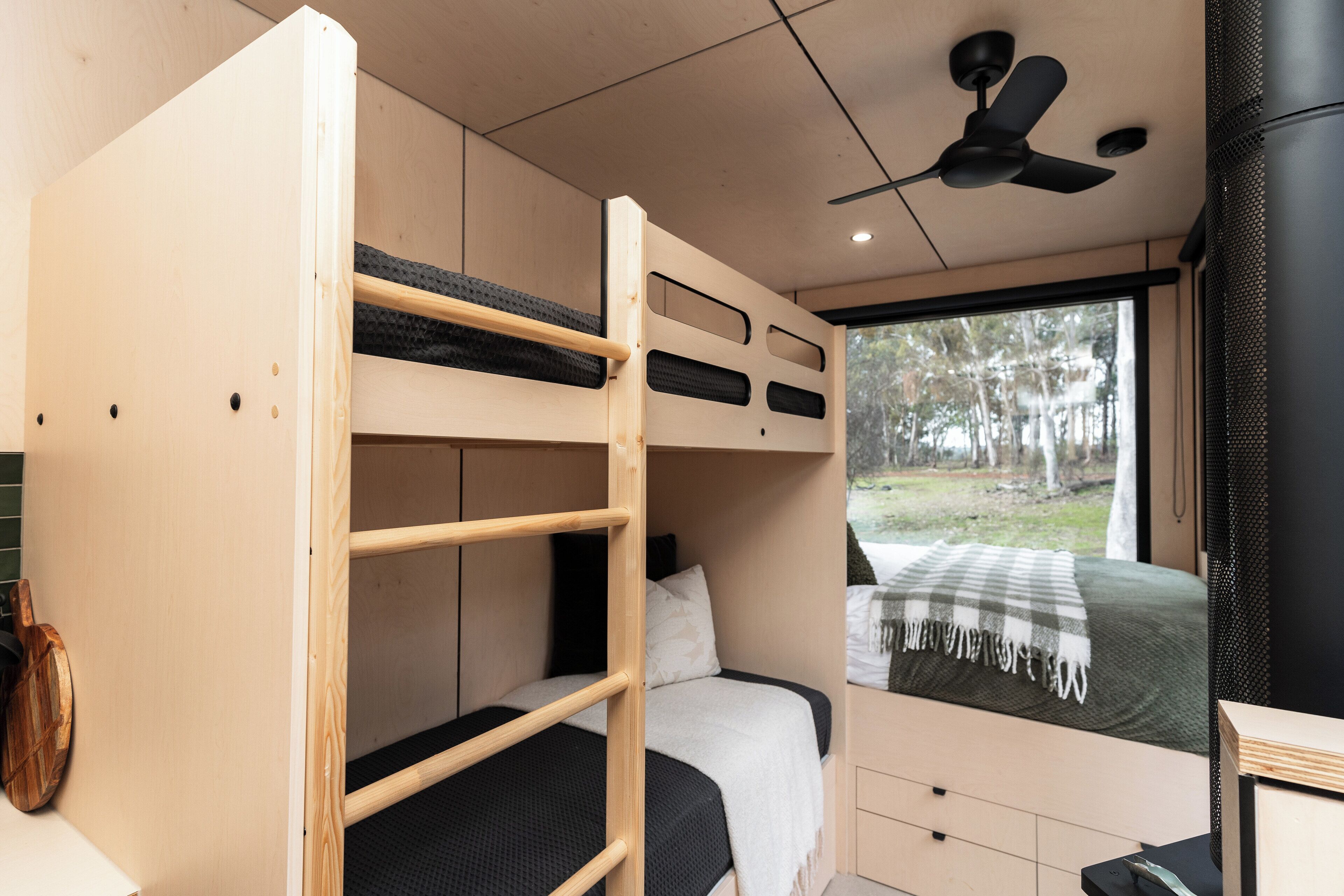 A bunk bed room with a ceiling fan, a window with a view of trees, and a small desk with a chair.