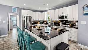 A modern kitchen with white cabinets, a large island with bar stools, stainless steel appliances, and a backsplash with black tiles. - Canal Front | Gated Community, Firepit & Decks (Port Bolivar)