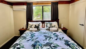 A bedroom with a bed, wooden headboard, two nightstands, a window with curtains, and an air conditioning unit. - Little Innes - affordable accommodation with a view (PORT ALBANY)