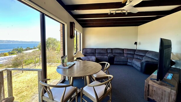 A modern living room with a large window, a round wooden table, and a dark-colored sectional sofa. - Little Innes - affordable accommodation with a view (PORT ALBANY)