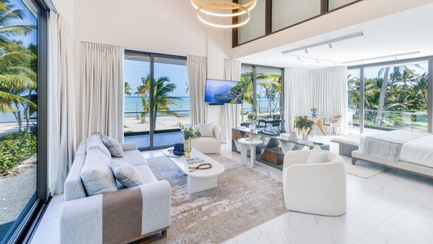 Un salon moderne avec un grand canapé, une table basse et un téléviseur. De grandes fenêtres sont agrémentées de rideaux, il y a également un coin repas et une vue sur l’océan.