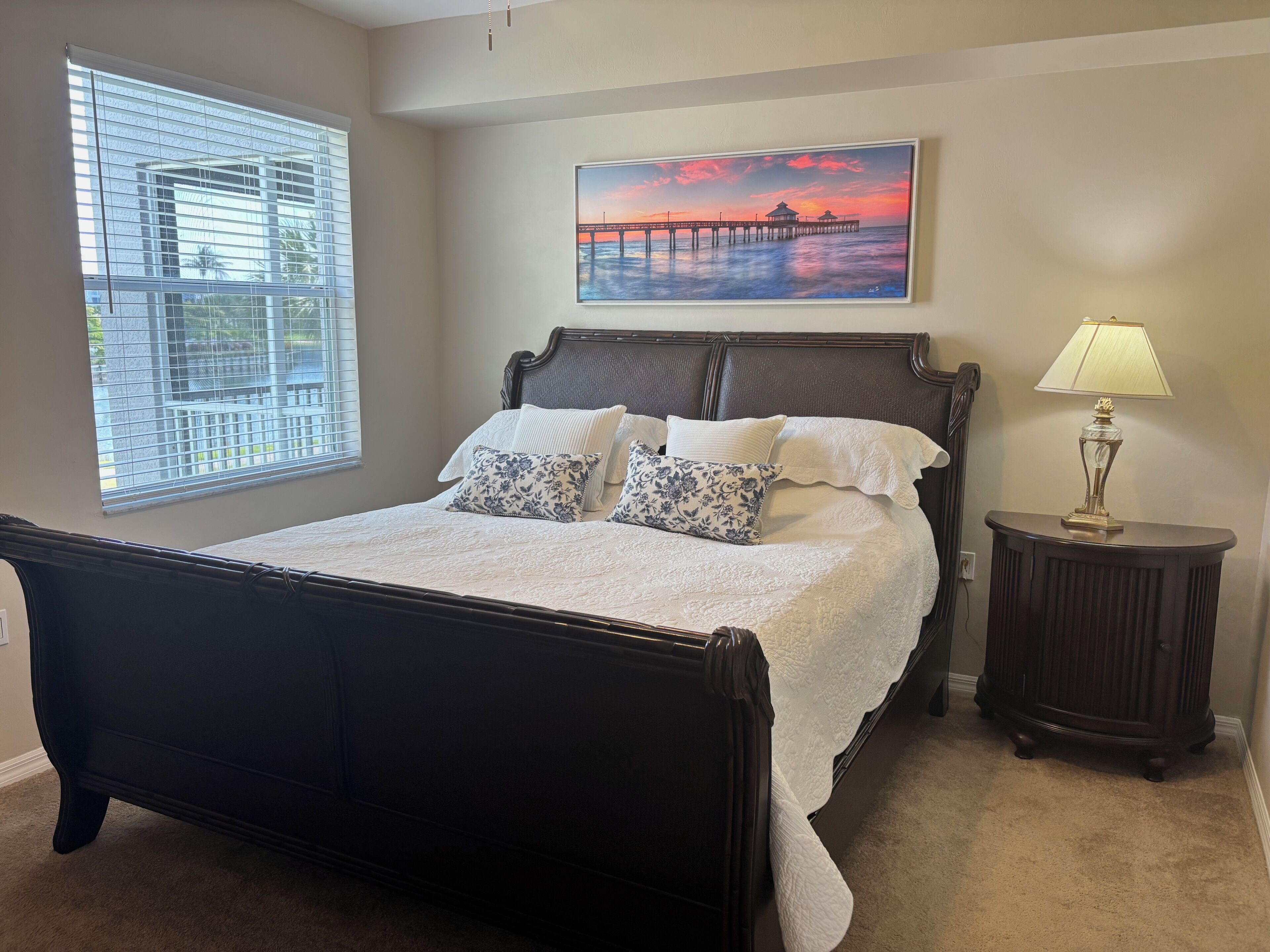 A bedroom with a bed, a nightstand, a lamp, a painting, and a window with blinds.