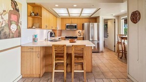 A modern kitchen with wooden cabinets, a central island with stools, a microwave, a refrigerator, and a dining area with chairs. - Beach front Honu Cottage (Kihei)