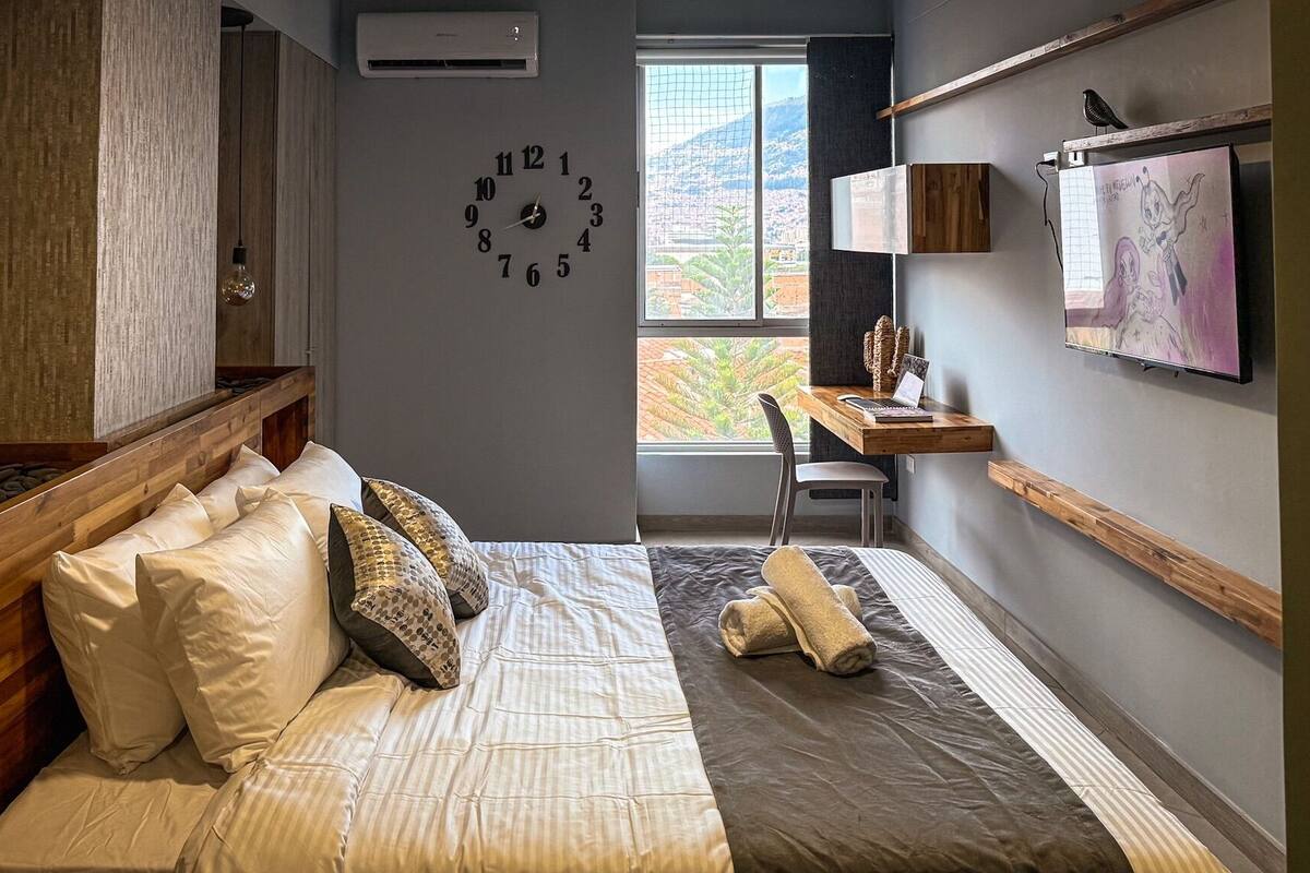 Ein Schlafzimmer mit Bett, Schreibtisch, Stuhl, einer Uhr und einem Fenster mit Blick auf eine Stadtlandschaft.