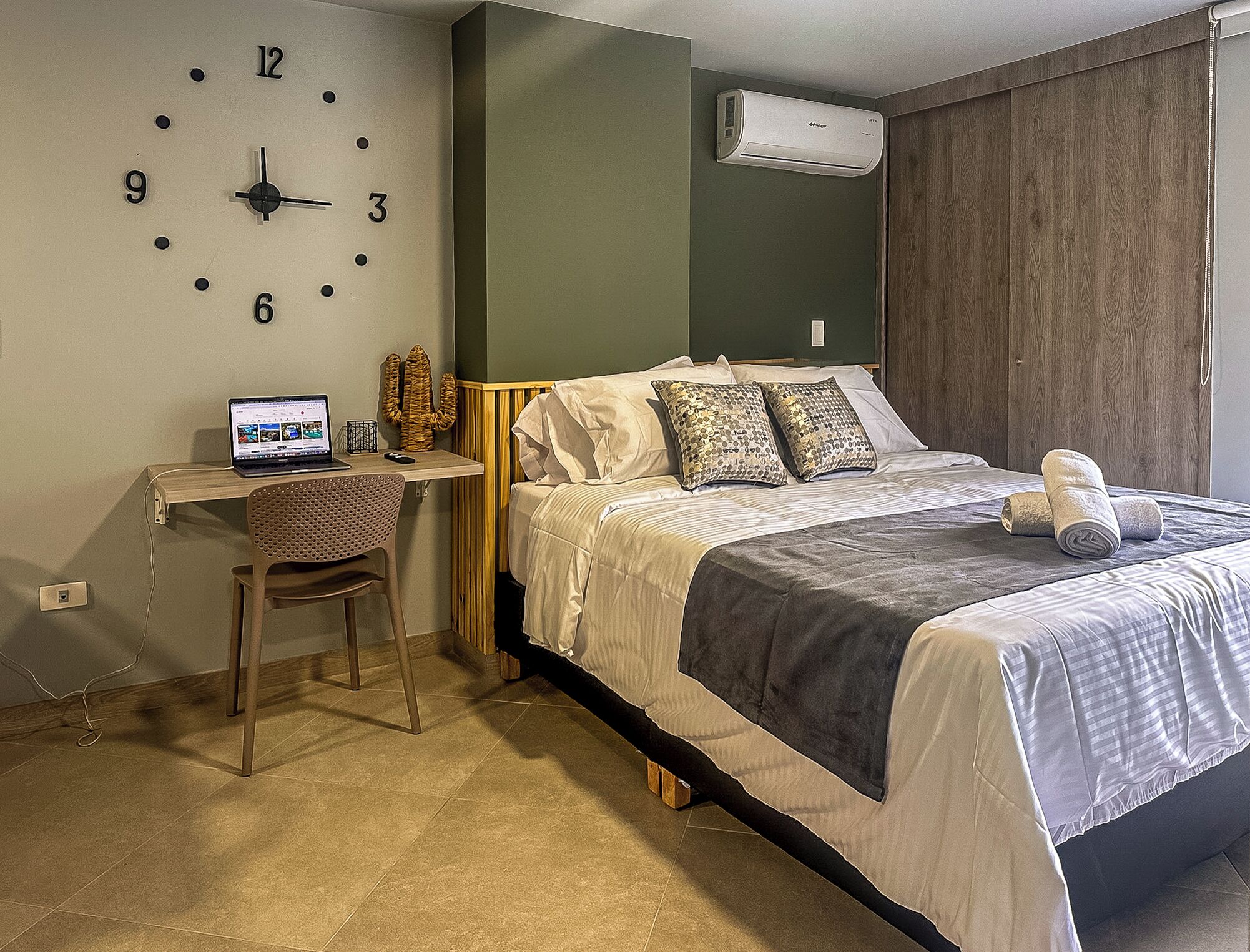 A bedroom with a bed, a desk with a laptop, a chair, a wall clock, and an air conditioner.