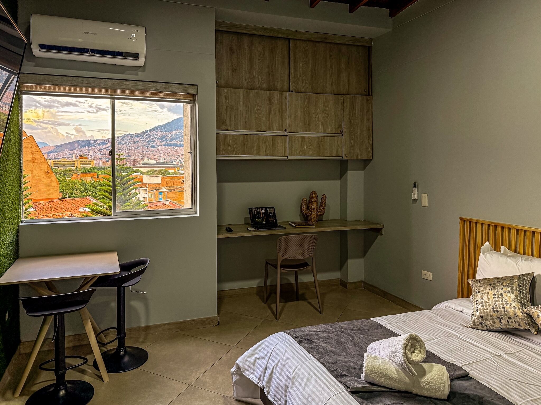 Ein Schlafzimmer mit Bett, Schreibtisch und Stuhl. Ein Fenster bietet Blick auf eine Stadtansicht.