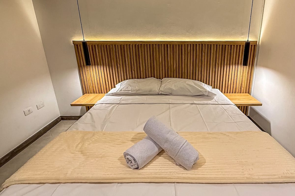 Ein ordentlich bezogenes Bett mit einem zusammengerollten Handtuch, einem Holz-Kopfteil und Nachttischen.
