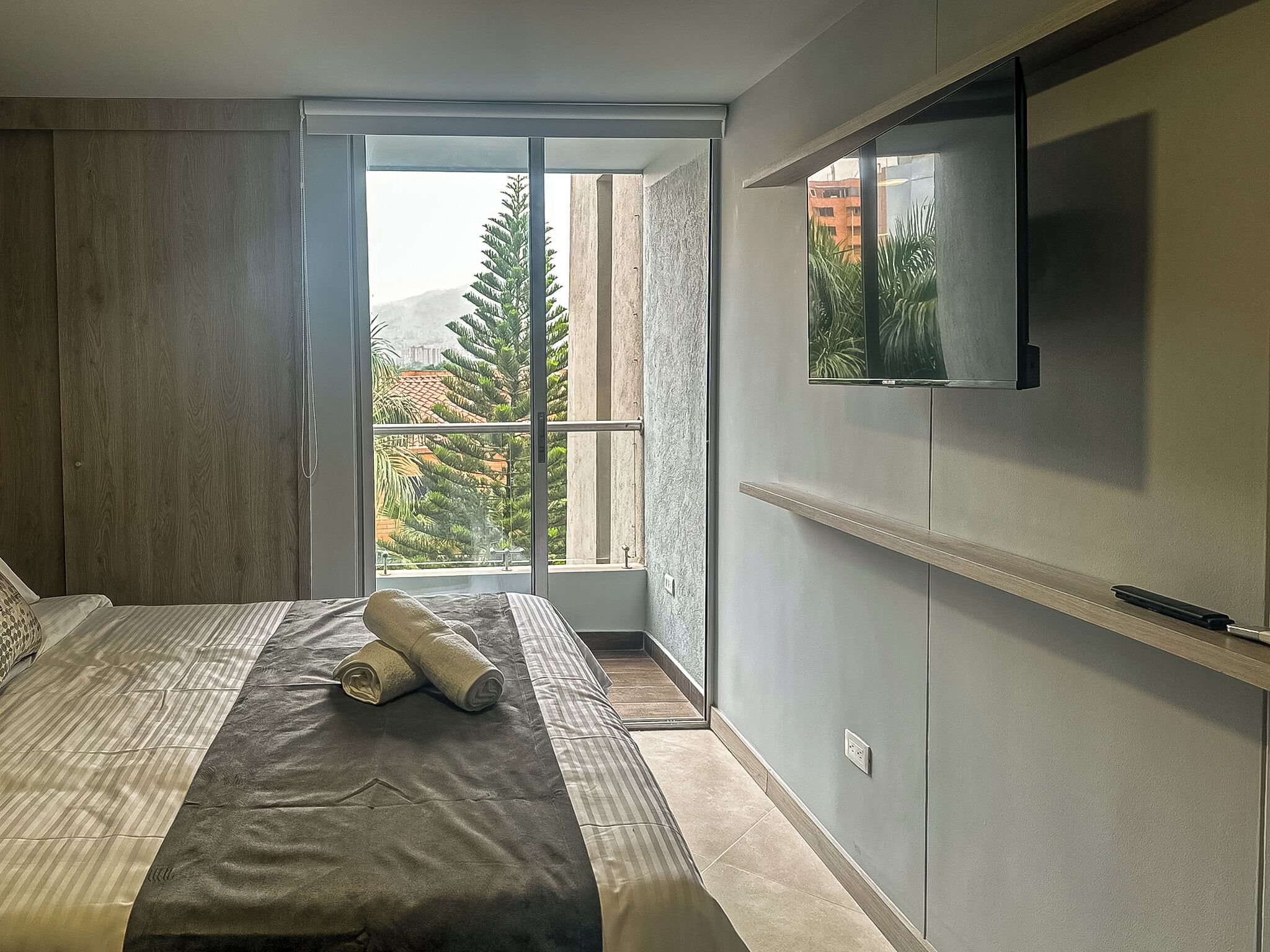 A modern bedroom with a large bed, a TV mounted on the wall, and a balcony with a view of buildings and trees.
