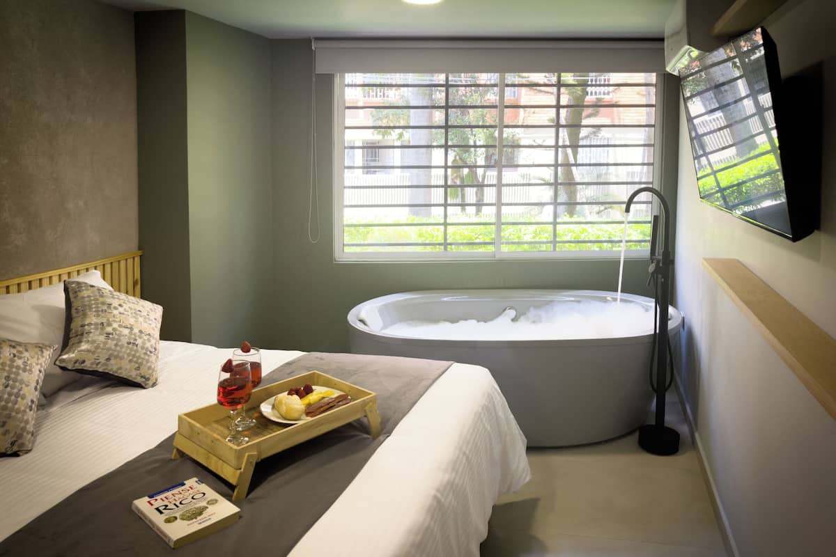 Ein Schlafzimmer mit einem großen Bett, einem Fenster mit Blick auf Grünflächen, einer Badewanne und einem Tablett mit Speisen und Getränken.