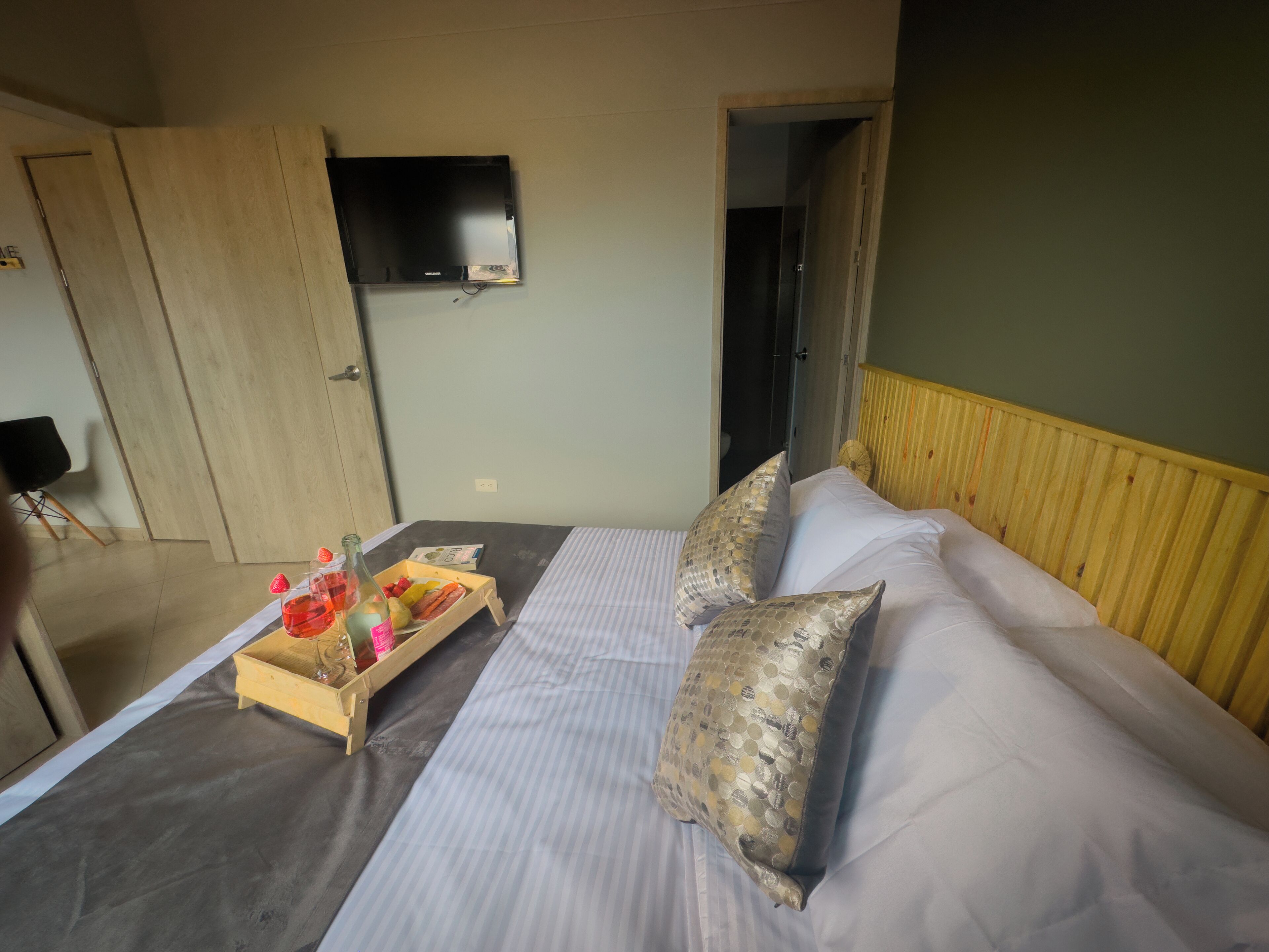 A hotel room with a bed, a wooden headboard, a television mounted on the wall, and a tray with fruits and a bottle on the bed.