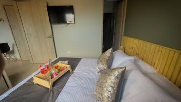 A hotel room with a bed, a wooden headboard, a television mounted on the wall, and a tray with fruits and a bottle on the bed.