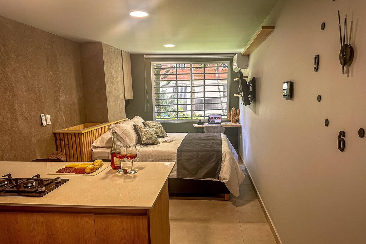 Ein kompaktes Hotelzimmer mit Küchenzeile, einem Bett mit Blick nach außen, einem kleinen Schreibtisch mit Laptop und einem an der Wand befestigten Fernseher.