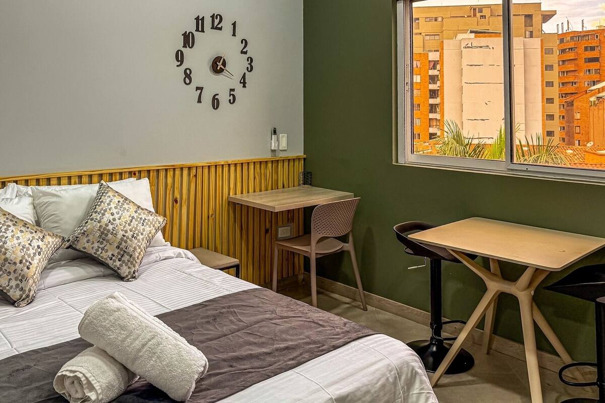 Ein Schlafzimmer mit Bett, Kissen, Nachttisch, Sessel, kleinem Tisch und Wanduhr.
