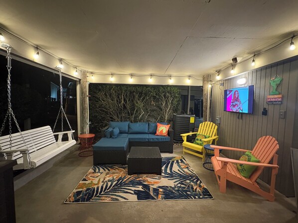 A covered outdoor seating area with a swing, sofas, chairs, and a TV screen.