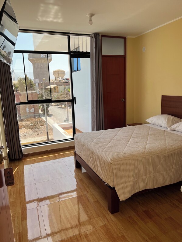 Une chambre d’hôtel avec un lit, un plancher en bois, une porte-fenêtre coulissante donnant sur un espace extérieur et une vue sur un château d’eau.