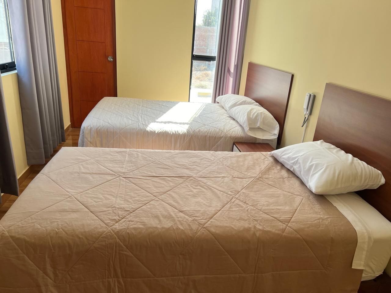 Quarto de hotel com duas camas, porta de madeira e janela com cortinas.