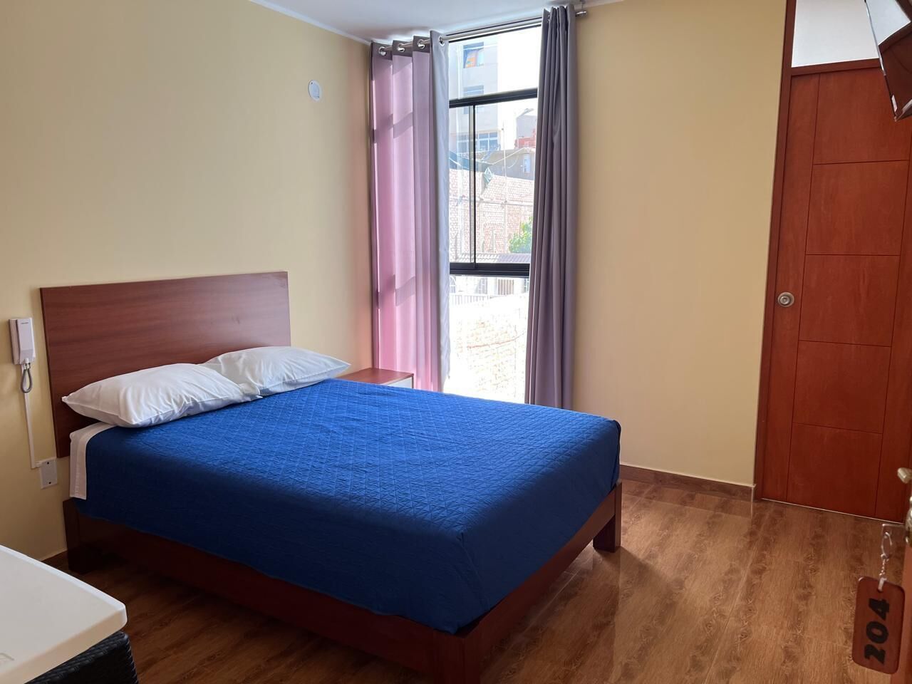 Une chambre d’hôtel avec un lit, une tête de lit en bois, une fenêtre avec des rideaux et une porte.