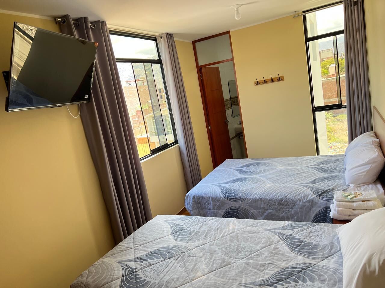 A room with two beds, a flat-screen TV mounted on the wall, and a window with curtains.